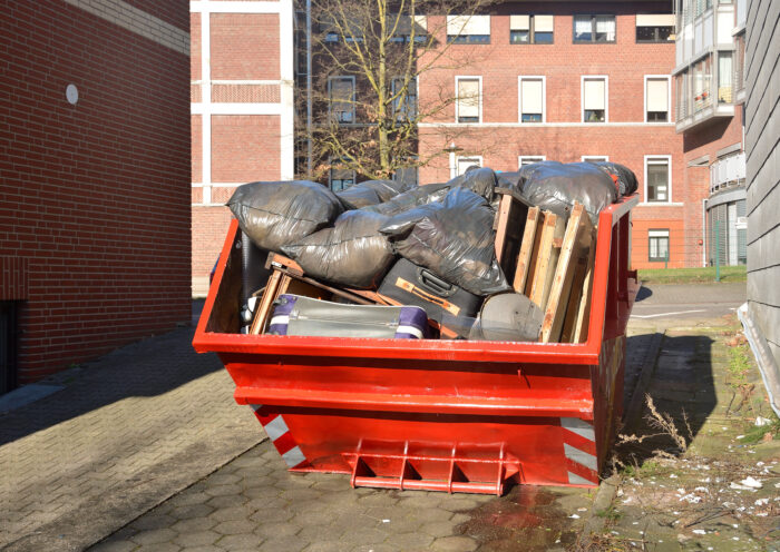 Full orange metal Skip container standing on the road near the house. In the container are old things, suitcases, furniture, black plastic bags of garbage and rubbish after cleaning the house.