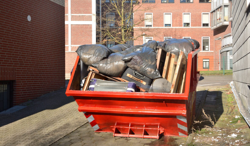 Full orange metal Skip container standing on the road near the house. In the container are old things, suitcases, furniture, black plastic bags of garbage and rubbish after cleaning the house.